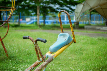 Rusty seesaw with blue seat in green park playground. A faded, rusted seesaw stands in a grassy park, its blue seat and yellow frame telling a story of long use.