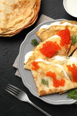 Tasty crepes with red caviar and sour cream on black textured table, flat lay