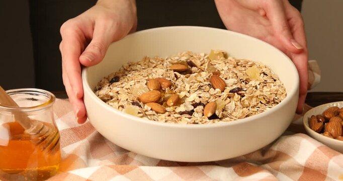 Making granola. Woman showing bowl of oat flakes, dried fruits and almond nuts at table against grey background, closeup