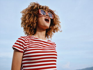 A joyful woman with curly hair wears a red and white striped tee and sunglasses, laughing under a clear blue sky, capturing a vibrant moment of sunshine and freedom.