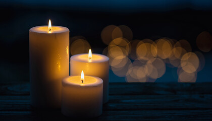 Three white candles cast a warm, tranquil glow on a dark wooden surface, with blurred golden bokeh lights. This serene scene evokes peace, comfort, and quiet contemplation