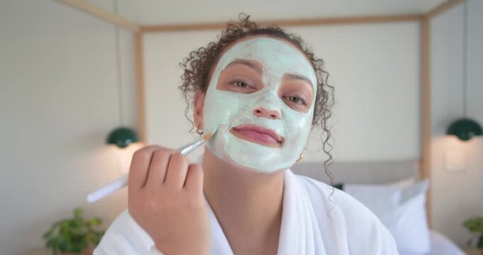 Woman in white robe picking brush, jar, smoothing green mask for skincare by four-poster bed