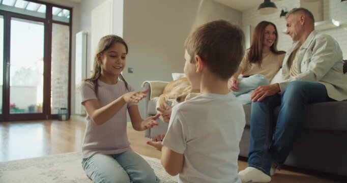 white siblings playing rock paper scissors on rug while parents watch