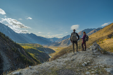 Fototapeta premium A hiking couple with backpacks and trekking poles stands on a mountain road edge, admiring a vast autumn valley below with a forest, river, and snow-capped peaks under a radiant blue sky with sunbeams