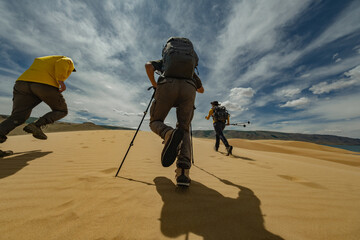 Hikers with backpacks ascend a Mongolian desert dune. Low rear-angle shot emphasizes boots and...
