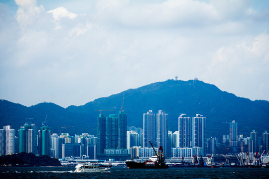 A coastal city with boats, cranes, high-rises, and forested mountains, showing urban growth and maritime activity.