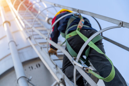 Close-up, low-angle shot of a construction worker in a safety harness climbing a cement silo ladder with sun flare, emphasizing safety protocols.