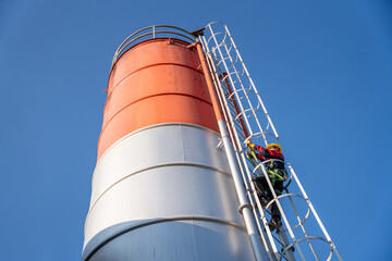 A construction worker in a safety harness climbs a tall, colorful cement silo ladder against a clear blue sky, emphasizing maintenance and safety.