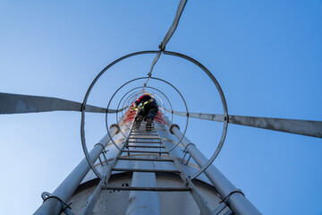 Close-up, low-angle shot of a construction worker in a yellow hard hat and safety harness climbing a cement silo ladder, looking down and ensuring safety.