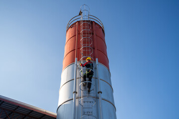 An extreme low-angle view of a worker climbing the fixed ladder of a tall cement silo, emphasizing industrial height and safety.