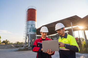 Two construction managers using a laptop for site inspection and planning at a small ready-mix concrete batching plant during sunset.