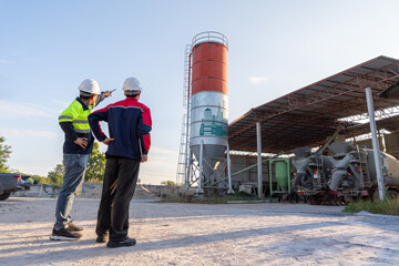 Two construction engineers stand watching a concrete mixer truck and a massive silo at a small-scale cement plant. Industrial management and planning.