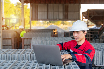 A construction manager uses a laptop to manage inventory, sitting among rows of fresh concrete cinder blocks at a small manufacturing plant.
