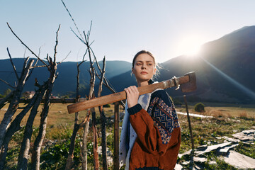 Obraz premium Woman carrying an axe on her shoulder in countryside near a rustic fence with mountains and sunrise in background, wearing a patterned sweater and looking confident.