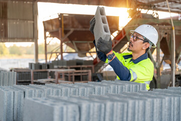 A construction manager carefully lifts a concrete cinder block for inspection, focusing on product quality at a small manufacturing plant during sunset.