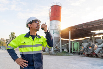 Construction manager using a two-way radio to coordinate production and operations at a small ready-mix concrete batching plant during sunset.