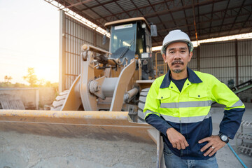 A confident construction manager in safety gear poses with a wheel loader behind him, overseeing industrial aggregate and machinery operations.