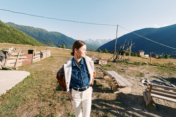 Obraz premium Woman in countryside with mountains and meadow, smiling outdoors while wearing a backpack and casual clothes. Rural landscape in sunlight, relaxed portrait and travel lifestyle scene.