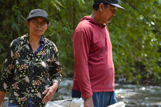 A man and woman stand near a shallow rocky river with bamboo behind them. The woman in a patterned top looks at the camera while the man in a red hooded shirt looks down. - Powered by Adobe