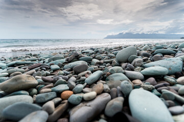 A close-up, low-angle shot of a beach covered in smooth, colorful, water-worn pebbles, with the ocean, waves, and a cloudy sky in the background.
