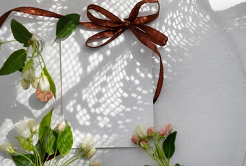 Blank card surrounded by delicate flowers and patterned shadows on a soft white surface in natural light