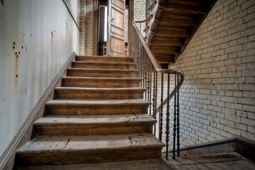Fototapeta premium An old, abandoned wooden staircase with a curving banister in a decaying building with brick walls, leading upwards to an open doorway.