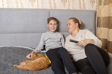 Happy Caucasian mother and preteen son relaxing together at home, sitting on the bed and watching...