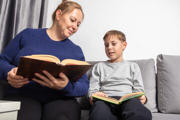 Caring mother helps her young Caucasian son read a book while sitting comfortably on a gray sofa at home, focusing on education and family bonding time.