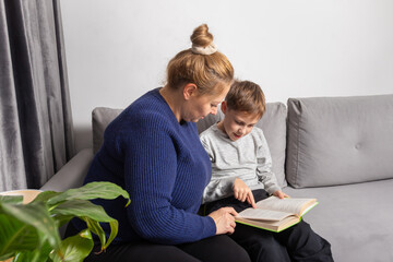 Caring mother helps her young Caucasian son read a book while sitting comfortably on a gray sofa at home, focusing on education and family bonding time.