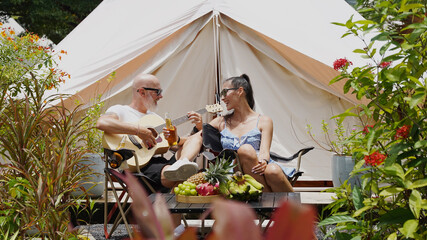 A man and woman is joyfully drinking beer and playing guitar near a cozy tent at glamping