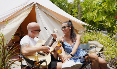A man and woman is joyfully drinking beer and playing guitar near a cozy tent at glamping