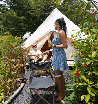 A man and woman is joyfully drinking beer and playing guitar near a cozy tent at glamping