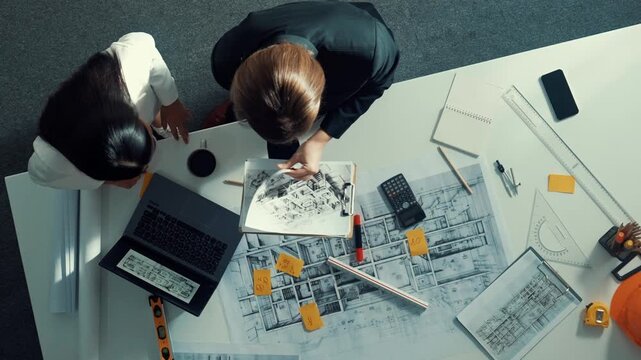 Top view of skilled project manager holding project plan and explain house design to architect engineer at meeting table with equipment, ruler, safety helmet and laptop scatter around. Alimentation.