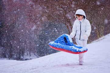 Happy Boy Walking Up Snowy Hill with Inflatable Sled - Winter Adventure in Falling Snow. Fun Seasonal Ride