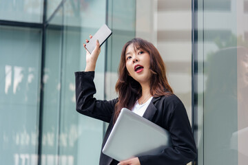 Young Asian business woman wearing suit, ready for work, holding laptop, raising phone with open mouth expression on busy city street, urban office building backdrop shows dynamic professional life