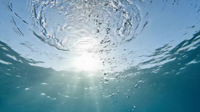 Underwater view looking up at sunlight rays piercing through blue ocean surface with rising bubbles, World Oceans Day concept, 4K