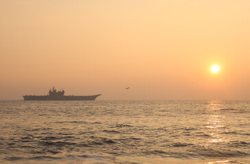 Trivandrum India December 3 2025 : Indian Navy aircraft carrier sails off Shangumugham Beach at golden sunset during Navy Day demo, with calm Arabian Sea reflecting warm light.