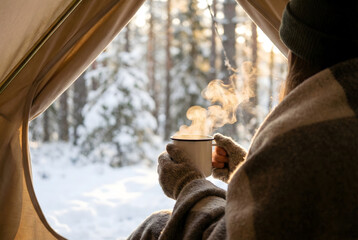 Person holding steaming mug inside tent looking at snowy forest winter camping