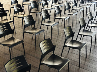 Empty black chairs arranged on a wooden floor in a spacious, well-lit room, creating a pattern of lines and shadows, evoking a sense of readiness for an event or meeting