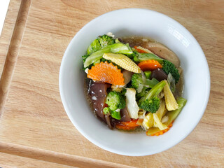 Colorful stir-fried mixed vegetables in oyster sauce, featuring broccoli, carrots, baby corn, and mushrooms, served in a white bowl on a wooden tray, fresh healthy plant-based cuisine
