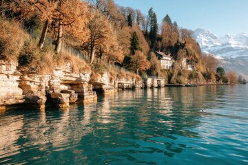 A serene lakeside scene featuring rocky cliffs, autumn foliage, and a quaint house against a backdrop of mountains and clear water.