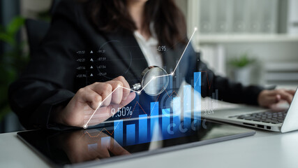 A businesswoman uses a tablet to analyze financial data with a magnifying glass. The modern office features graphs and a laptop, showcasing a professional work environment. Scalp