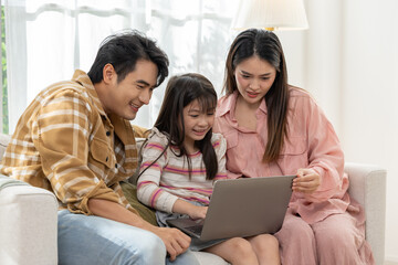 Asian happy family watching laptop together on sofa sharing joyful moment of bonding and learning with child smiling and parents attentive in bright living room