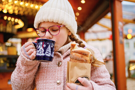 Little cute school girl eating sweet bread called Baumstriezel and drink hot chocolate or punch on German Christmas market. Happy child on traditional family market in Germany,