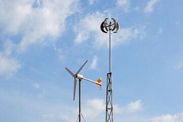Two types of wind turbines are installed against a cloudy blue sky: a horizontal-axis turbine and a vertical Savonius-style cup anemometer, illustrating different wind power technologies.