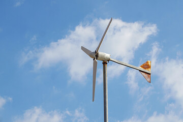 A small, horizontal-axis wind turbine with three blades and a tail vane stands against a bright, cloudy blue sky, representing personal or small-scale wind power generation.