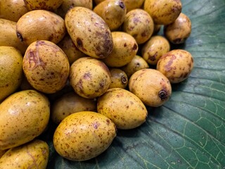 Close-up of a pile of small, yellowish-brown, speckled tropical fruits (hog plums or Spondias dulcis) lying on a large green leaf.