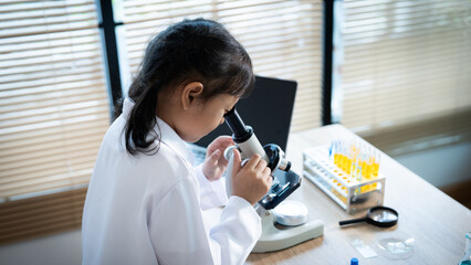 A young girl is looking through a microscope at a slide. She is wearing a white lab coat and she is...
