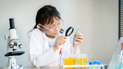 A young girl wearing a lab coat and glasses is looking through a magnifying glass at a small object