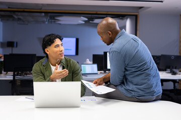 Diverse male coworkers discussing project data in open-plan office with laptop and printed charts